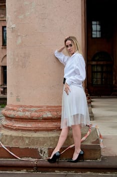 Young blonde woman dreses in white skirt and shirt posing on the steps of the old looking vintage building with large 
columns and bas-reliefs. Fashion look's woman. Young woman's modern portrait.