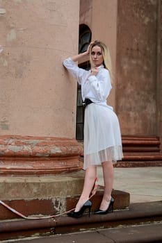 Young blonde woman dreses in white skirt and shirt posing on the steps of the old looking vintage building with large 
columns and bas-reliefs. Fashion look's woman. Young woman's modern portrait.