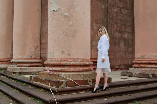 Young blonde woman dreses in white skirt and shirt posing on the steps of the old looking vintage building with large 
columns and bas-reliefs. Fashion look's woman. Young woman's modern portrait.