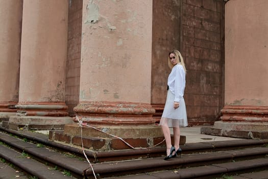 Young blonde woman dreses in white skirt and shirt posing on the steps of the old looking vintage building with large 
columns and bas-reliefs. Fashion look's woman. Young woman's modern portrait.