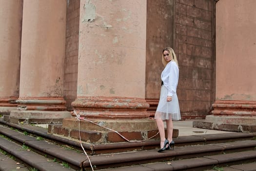 Young blonde woman dreses in white skirt and shirt posing on the steps of the old looking vintage building with large 
columns and bas-reliefs. Fashion look's woman. Young woman's modern portrait.