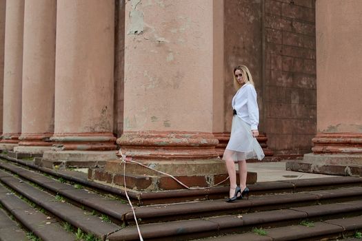 Young blonde woman dreses in white skirt and shirt posing on the steps of the old looking vintage building with large 
columns and bas-reliefs. Fashion look's woman. Young woman's modern portrait.