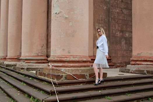 Young blonde woman dreses in white skirt and shirt posing on the steps of the old looking vintage building with large 
columns and bas-reliefs. Fashion look's woman. Young woman's modern portrait.