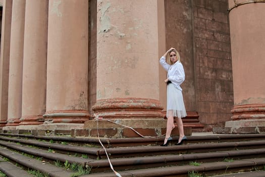 Young blonde woman dreses in white skirt and shirt posing on the steps of the old looking vintage building with large 
columns and bas-reliefs. Fashion look's woman. Young woman's modern portrait.