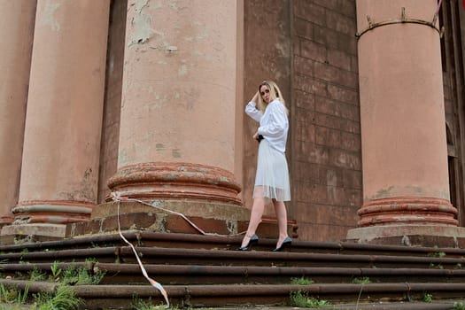 Young blonde woman dreses in white skirt and shirt posing on the steps of the old looking vintage building with large 
columns and bas-reliefs. Fashion look's woman. Young woman's modern portrait.