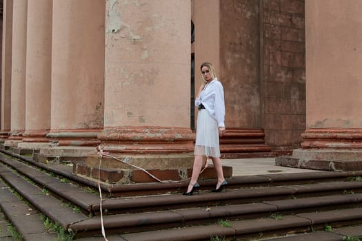 Young blonde woman dreses in white skirt and shirt posing on the steps of the old looking vintage building with large 
columns and bas-reliefs. Fashion look's woman. Young woman's modern portrait.