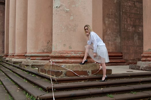 Young blonde woman dreses in white skirt and shirt posing on the steps of the old looking vintage building with large 
columns and bas-reliefs. Fashion look's woman. Young woman's modern portrait.