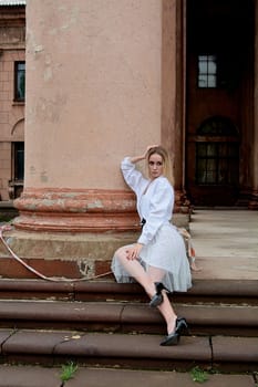 Young blonde woman dreses in white skirt and shirt posing on the steps of the old looking vintage building with large 
columns and bas-reliefs. Fashion look's woman. Young woman's modern portrait.