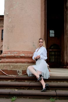 Young blonde woman dreses in white skirt and shirt posing on the steps of the old looking vintage building with large 
columns and bas-reliefs. Fashion look's woman. Young woman's modern portrait.