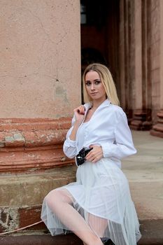 Young blonde woman dreses in white skirt and shirt posing on the steps of the old looking vintage building with large 
columns and bas-reliefs. Fashion look's woman. Young woman's modern portrait.