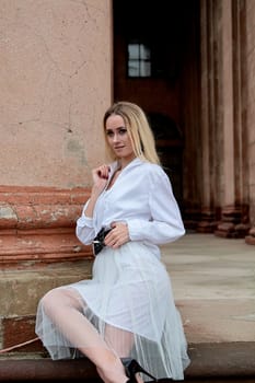 Young blonde woman dreses in white skirt and shirt posing on the steps of the old looking vintage building with large 
columns and bas-reliefs. Fashion look's woman. Young woman's modern portrait.