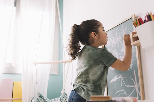 little girl focused drawing with a chalk on the blackboard at her room at home, copy space for text