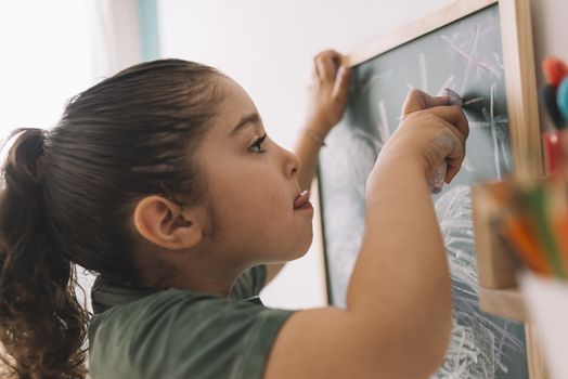 little girl focused drawing with a chalk on the blackboard at her room at home, copy space for text