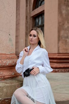 Young blonde woman dreses in white skirt and shirt posing on the steps of the old looking vintage building with large 
columns and bas-reliefs. Fashion look's woman. Young woman's modern portrait.