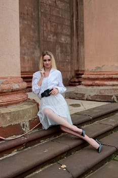 Young blonde woman dreses in white skirt and shirt posing on the steps of the old looking vintage building with large 
columns and bas-reliefs. Fashion look's woman. Young woman's modern portrait.