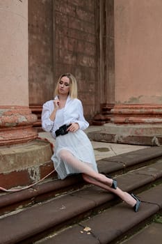 Young blonde woman dreses in white skirt and shirt posing on the steps of the old looking vintage building with large 
columns and bas-reliefs. Fashion look's woman. Young woman's modern portrait.