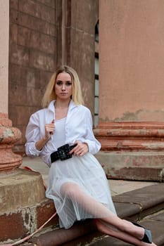 Young blonde woman dreses in white skirt and shirt posing on the steps of the old looking vintage building with large 
columns and bas-reliefs. Fashion look's woman. Young woman's modern portrait.