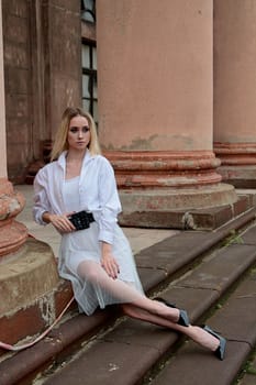 Young blonde woman dreses in white skirt and shirt posing on the steps of the old looking vintage building with large 
columns and bas-reliefs. Fashion look's woman. Young woman's modern portrait.