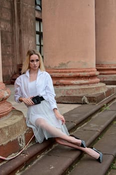 Young blonde woman dreses in white skirt and shirt posing on the steps of the old looking vintage building with large 
columns and bas-reliefs. Fashion look's woman. Young woman's modern portrait.