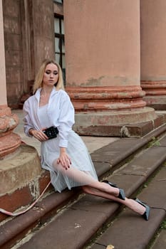 Young blonde woman dreses in white skirt and shirt posing on the steps of the old looking vintage building with large 
columns and bas-reliefs. Fashion look's woman. Young woman's modern portrait.