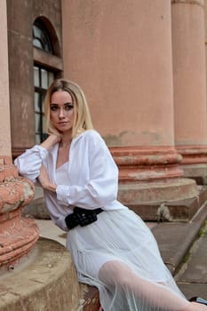 Young blonde woman dreses in white skirt and shirt posing on the steps of the old looking vintage building with large 
columns and bas-reliefs. Fashion look's woman. Young woman's modern portrait.