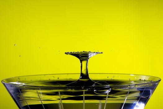 Droplets collide above martini glass, creating a radial fan pattern. Background is yellow.