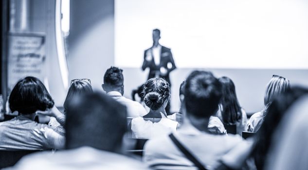 Speaker giving talk at business event. Audience at the conference hall. Business and Entrepreneurship concept. Focus on unrecognizable people in audience. Blue toned grayscale image.