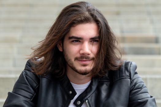 Portrait of a handsome bearded man with long brown hair, dressed in trendy clothes, sitting on the steps and looking to the side, with area for advertising content. Handsome hipster guy posing on copy space.