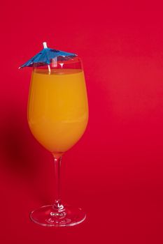 Orange drink with a blue umbrella in a wine glass set against a solid red background.