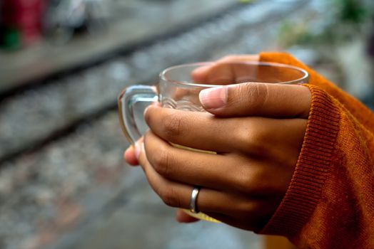 holding a cup of tea by rails waiting for the train with orange pullover covering hands by a third