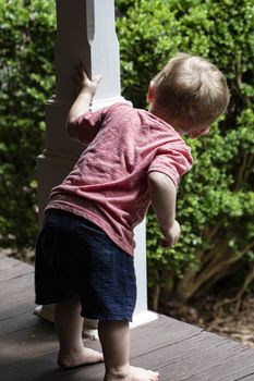 Barefoot male toddler peers around post on wooden porch.