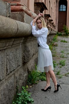 Young blonde woman in white skirt and shirt near the wall of the old looking vintage building. Fashion woman. Young woman's modern portrait.