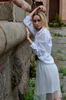 Young blonde woman in white skirt and shirt near the wall of the old looking vintage building. Fashion woman. Young woman's modern portrait.