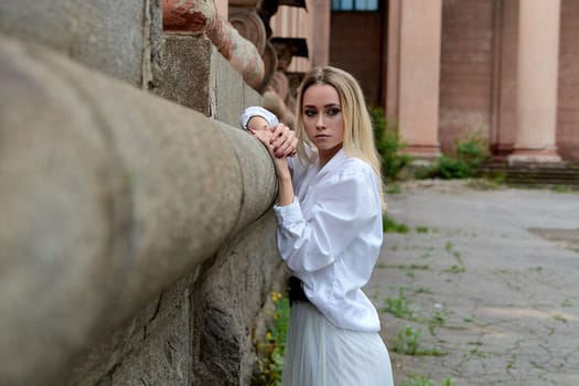 Young blonde woman in white skirt and shirt near the wall of the old looking vintage building. Fashion woman. Young woman's modern portrait.