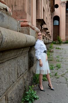 Young blonde woman in white skirt and shirt near the wall of the old looking vintage building. Fashion woman. Young woman's modern portrait.