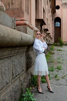 Young blonde woman in white skirt and shirt near the wall of the old looking vintage building. Fashion woman. Young woman's modern portrait.