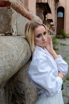 Young blonde woman in white skirt and shirt near the wall of the old looking vintage building. Fashion woman. Young woman's modern portrait.