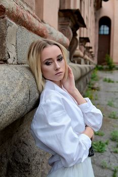 Young blonde woman in white skirt and shirt near the wall of the old looking vintage building. Fashion woman. Young woman's modern portrait.