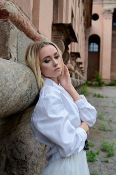 Young blonde woman in white skirt and shirt near the wall of the old looking vintage building. Fashion woman. Young woman's modern portrait.