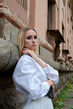Young blonde woman in white skirt and shirt near the wall of the old looking vintage building. Fashion woman. Young woman's modern portrait.