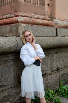 Young blonde woman in white skirt and shirt near the wall of the old looking vintage building. Fashion woman. Young woman's modern portrait.