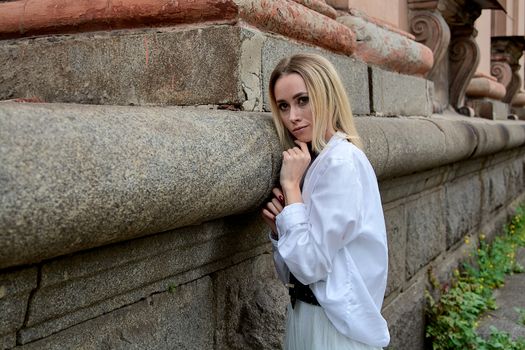 Young blonde woman in white skirt and shirt near the wall of the old looking vintage building. Fashion woman. Young woman's modern portrait.