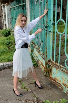 Young blonde woman in white skirt and shirt near the green vintage gate. Young woman's modern portrait.