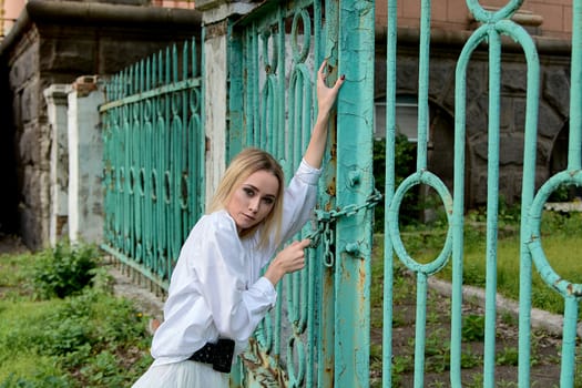 Young blonde woman in white skirt and shirt near the green vintage gate. Young woman's modern portrait.