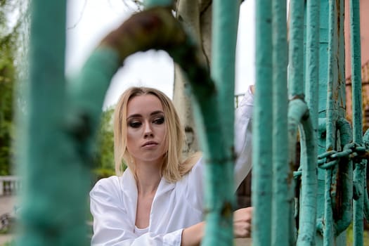 Young blonde woman in white skirt and shirt near the green vintage gate. Young woman's modern portrait.