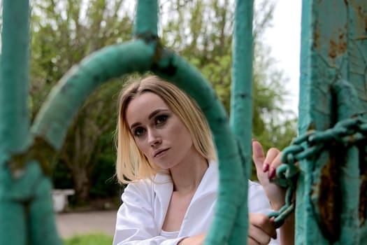 Young blonde woman in white skirt and shirt near the green vintage gate. Young woman's modern portrait.