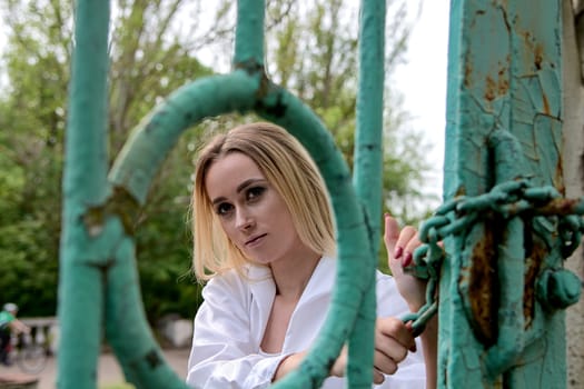 Young blonde woman in white skirt and shirt near the green vintage gate. Young woman's modern portrait.