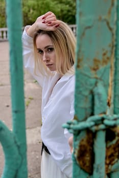 Young blonde woman in white skirt and shirt near the green vintage gate. Young woman's modern portrait.
