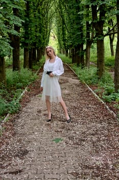 Young blonde woman in white skirt and shirt walks alone along the chestnut alley in the city park. Fashion woman. Young woman's modern portrait.