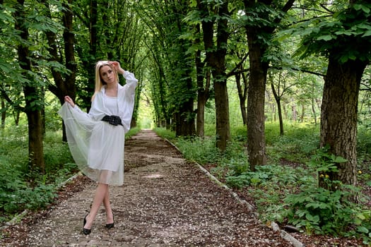 Young blonde woman in white skirt and shirt walks alone along the chestnut alley in the city park. Fashion woman. Young woman's modern portrait.